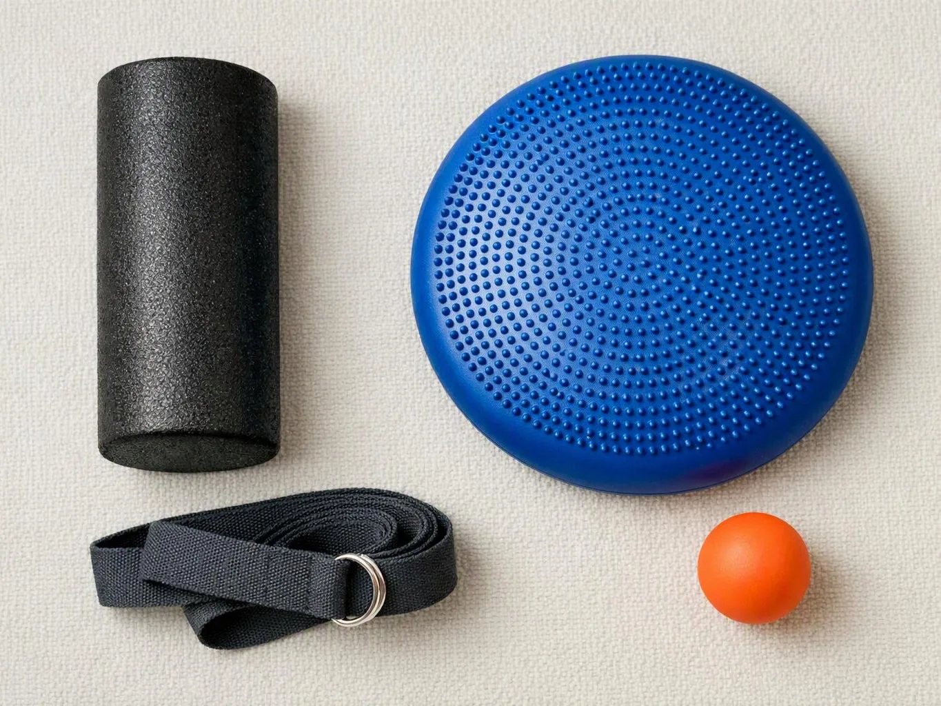 A black foam roller, blue textured balance disc, black strap, and orange exercise ball rest on a light carpet