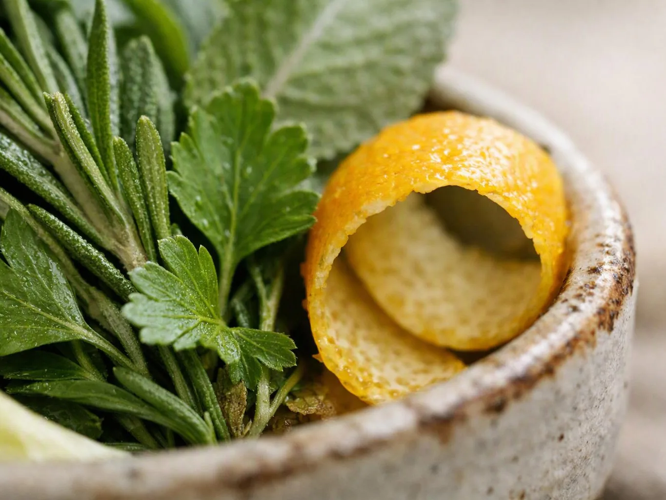 Fresh herbs and a curled orange peel sit in a rustic ceramic bowl