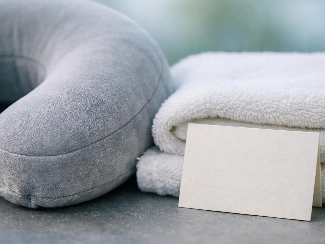 A gray neck pillow sits beside folded white towels with a blank card on a dark surface