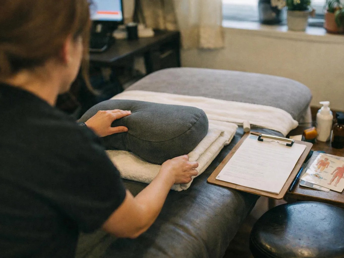 A person arranges a gray cushion on a massage table beside a clipboard in a calm treatment room