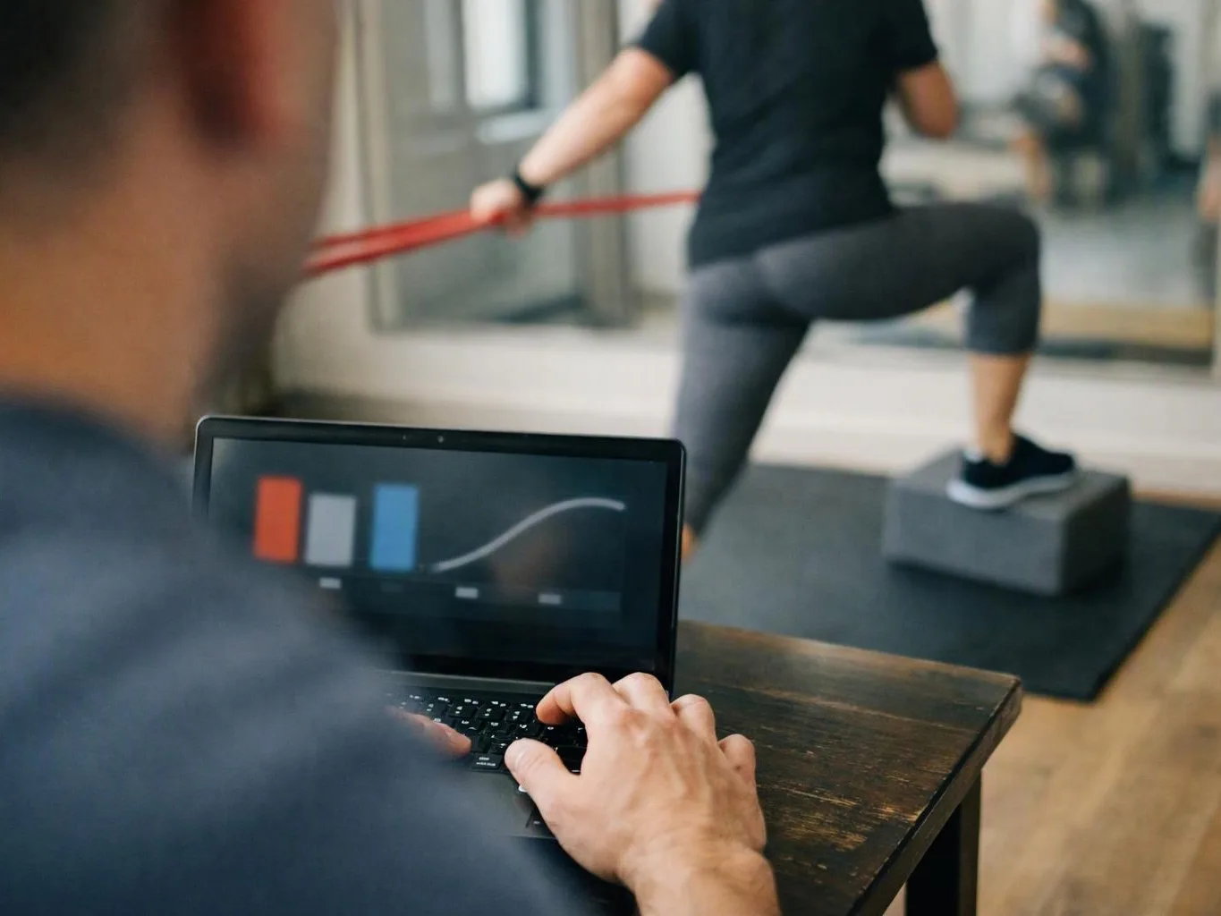 A person types on a laptop while another person exercises with a red resistance band in a gym studio