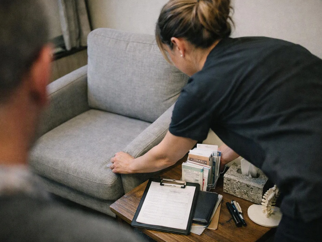 A woman adjusts a gray sofa beside a wooden side table with papers, pens, and tissue box